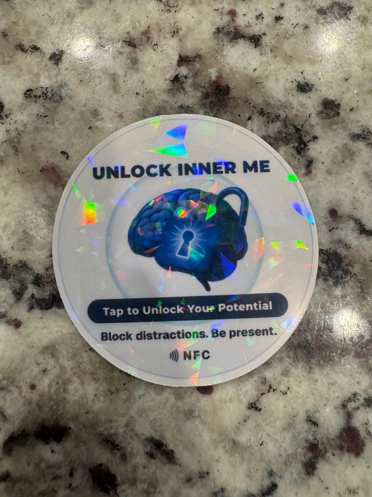 A round holographic NFC sticker. The sticker features a blue brain and padlock graphic with the text 'UNLOCK INNER ME', 'Tap to Unlock Your Potential', and 'NFC' printed at the bottom.